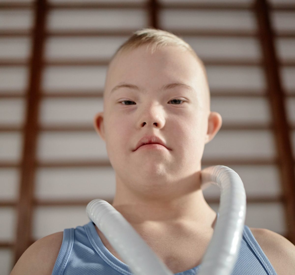 Portrait of Caucasian boy child with Down syndrome looking into camera, wearing sportswear, standing indoors in gym setting, short blond hair, neutral facial expression